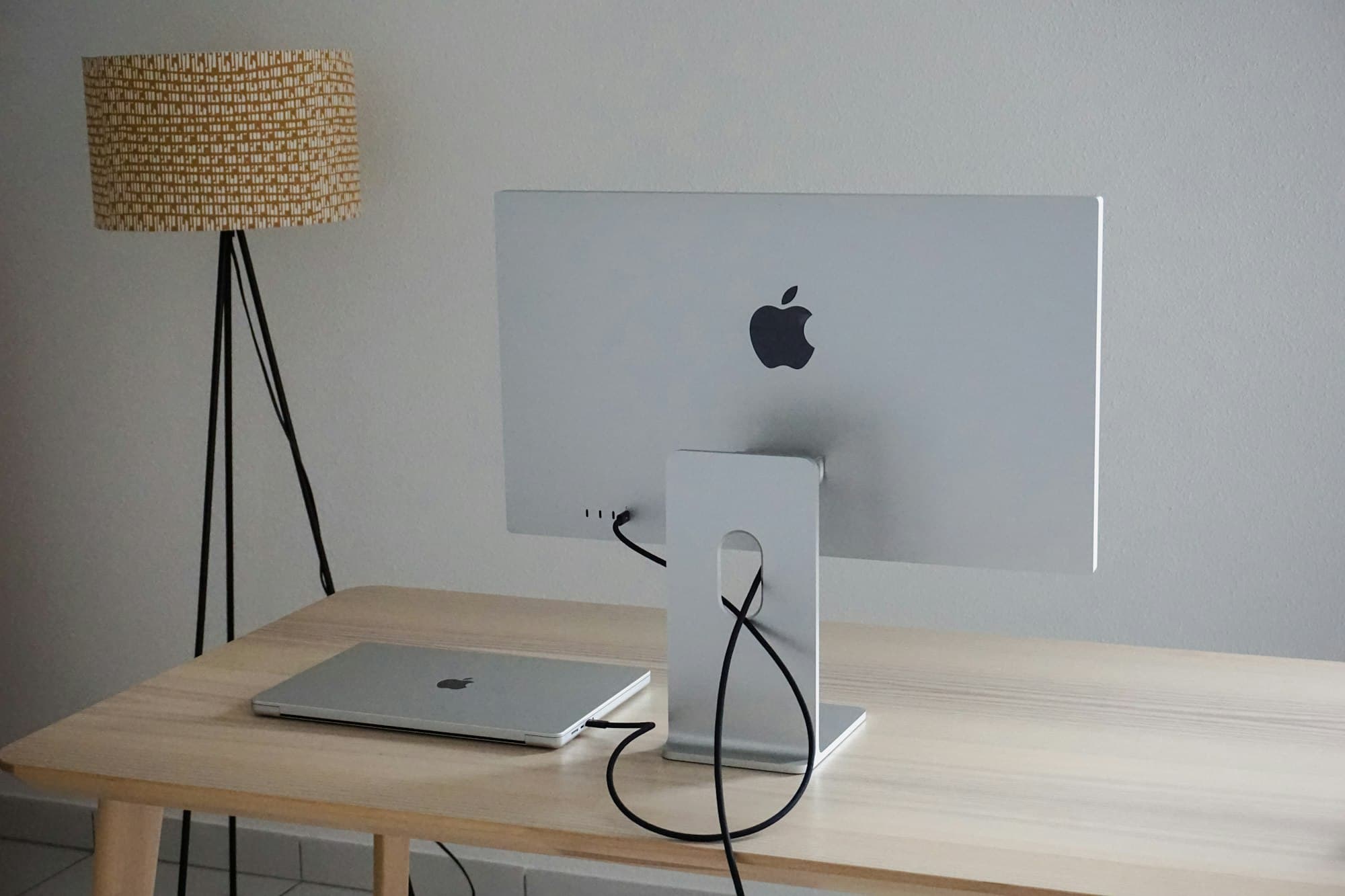 The back of an Apple Studio Display on a wooden desk, its cable running through the stand to a closed laptop